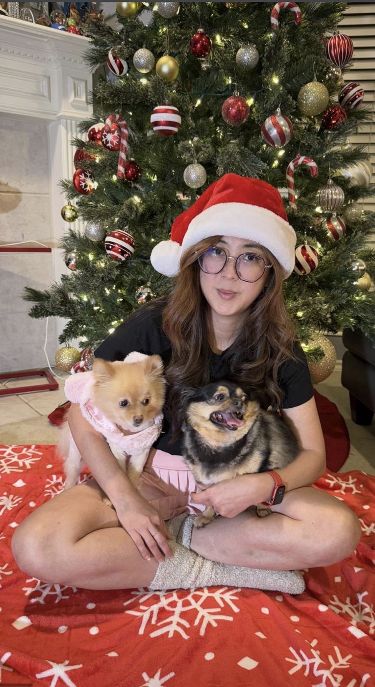 Christine in a Santa hat holding two small dogs in front of a Christmas tree
